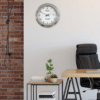 Modern office corner with a black leather chair, a wooden table with black metal legs, a small side table holding potted plants and books, a brick wall on the left, and a Rolex-style wall clock showing 10:11:50 with a date display on a plain white wall.