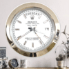 Large wall clock designed to resemble a Rolex Oyster Perpetual Day-Date watch with a gold rim, white face, rectangular hour markers, and displaying Martedì the 17th, mounted on a light-colored wall above a shelf with a small clock, a mug, decorative boxes, and a potted plant under a glass dome.