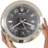 Hands holding a large round wall clock designed like a Rolex Oyster Perpetual Datejust watch with a black face, silver Roman numerals, and a date display showing 29.