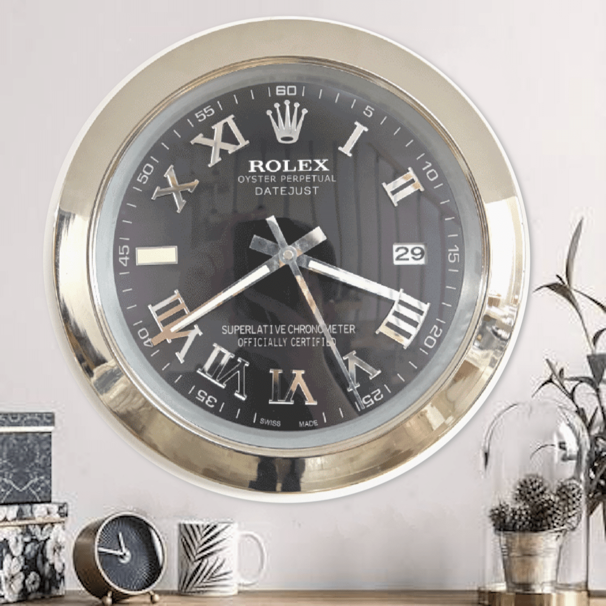 Wall-mounted Rolex Oyster Perpetual Datejust clock with Roman numerals, gold rim, showing time 8:16, above a wooden surface with decorative objects including a small clock, a patterned mug, and a glass dome with pine cones and a plant on the right.