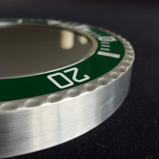 Close-up of a metallic watch bezel with a green insert marked with white numbers and indicators, showing the number 20 prominently, on a dark background.