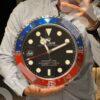 Person holding a large Rolex wall clock designed like a wristwatch with a black dial, blue and red bezel, bold white markers, and a date window showing 30.