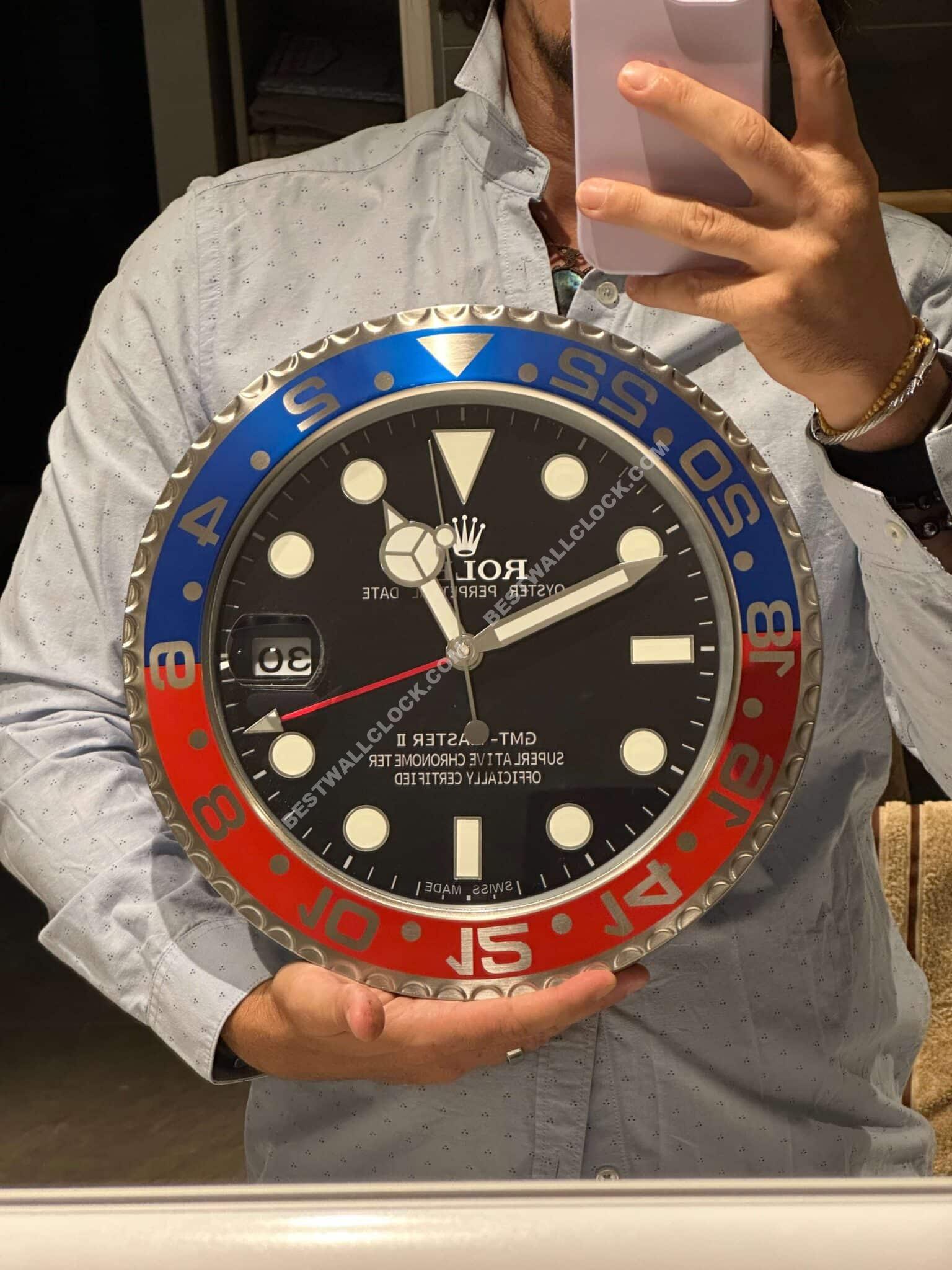 Person holding a large Rolex wall clock designed like a wristwatch with a black dial, blue and red bezel, bold white markers, and a date window showing 30.